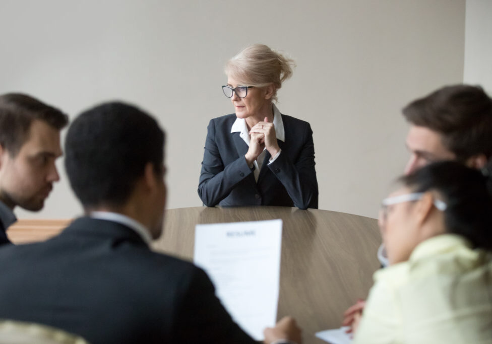 Middle aged stressed woman sitting at the desk in office at meeting. Young multiracial workers team sitting their backs to camera. Passing interview, hiring, recruiting and age discrimination concept