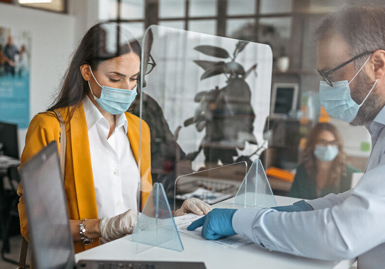 Bank teller discussing paperwork with customer at bank counter wearing protective gloves and face mask. Office with acrylic glass partition on desk. Acrylic glass wall - protection against coughs and spitting, protection against viruses.